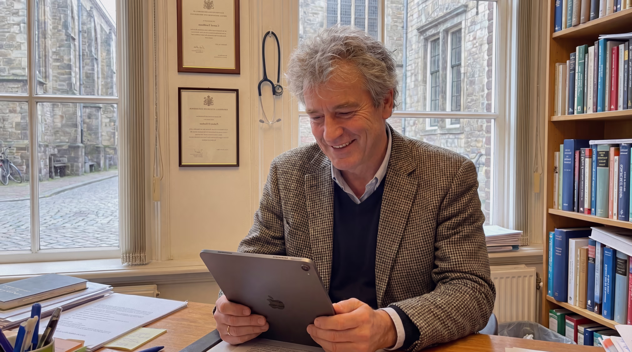 A grey-haired doctor in a tweed jacket smiles while reading a tablet in his office